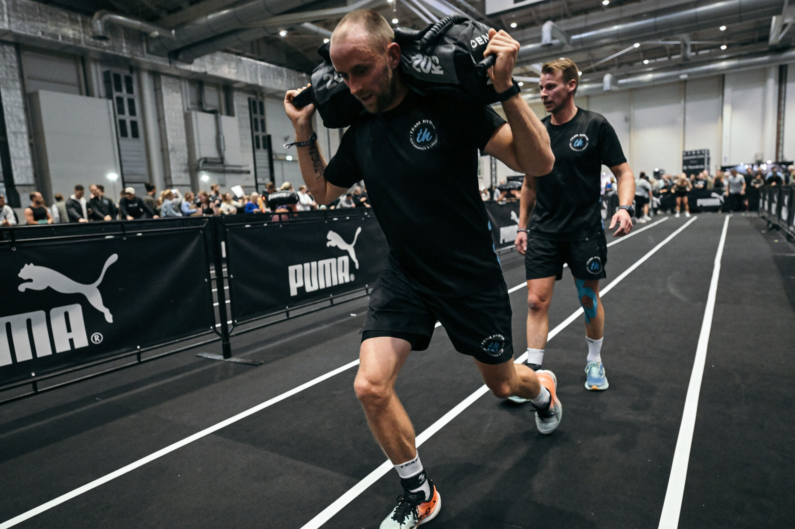 TEAM-HYBRID-Athlet mit Sandbag auf den Schultern auf der Laufstrecke, ein Teamkollege begleitet ihn, beim HYROX Hamburg 2025